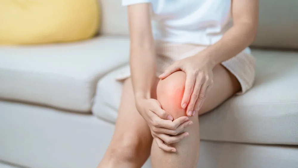 A woman sitting on the edge of a sofa at home, holding a stiff knee in the first minutes after getting up